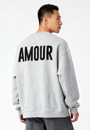 Jonge man zijwaarts kijkend, draagt een grijze trui met "AMOUR" in grote zwarte letters op de rug gedrukt.