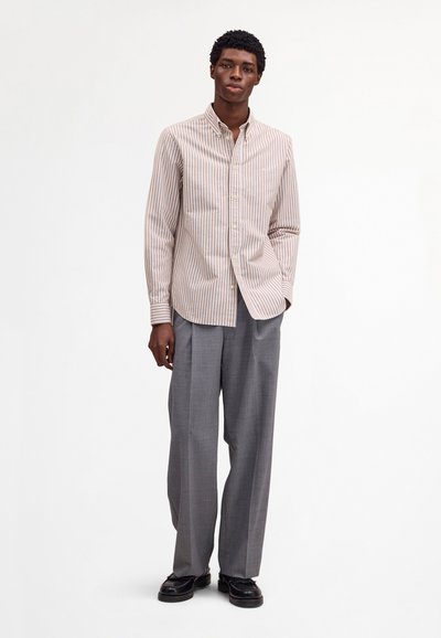 Młody mężczyzna stoi, ubrany w beżowo-białą koszulę w paski z guzikami, szare szerokie spodnie i czarne eleganckie buty na białym tle.