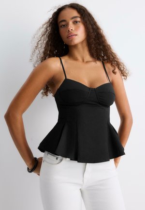 Femme aux cheveux bouclés portant un haut noir moulant sans manches avec de fines bretelles et un pantalon blanc, posant devant un fond uni.