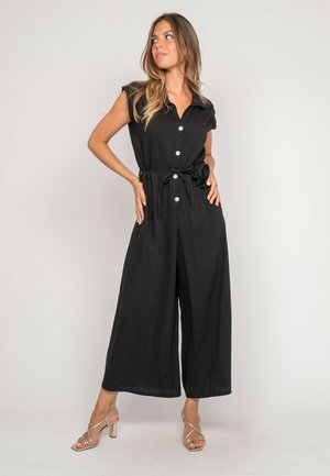 Femme portant une combinaison noire sans manches avec des boutons et une ceinture nouée, debout les mains sur les hanches, regardant sur le côté, sandales beiges à talons.