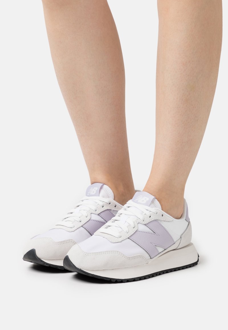 Paire de baskets New Balance blanches et violet clair portées pieds nus sur un fond blanc uni.