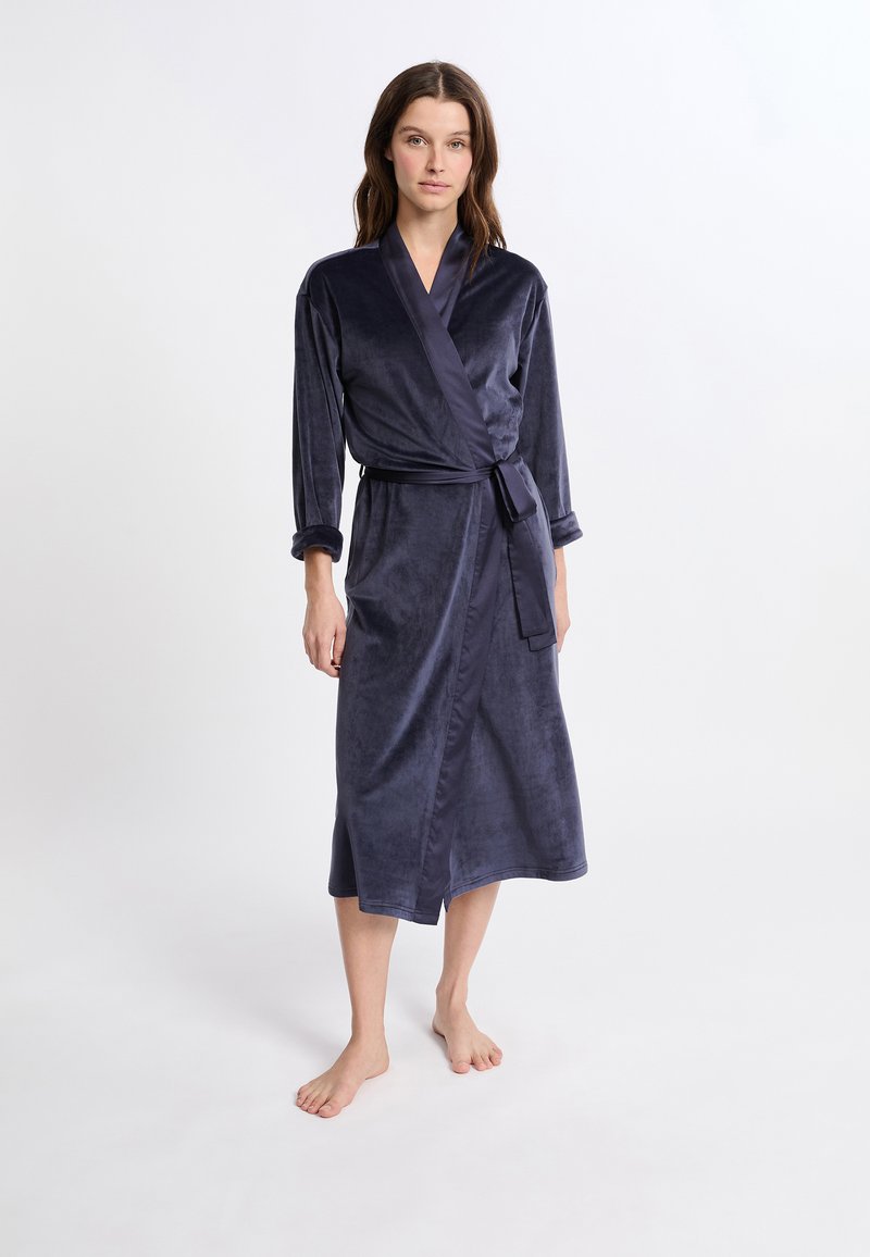 Femme debout pieds nus, portant une longue robe en velours bleu foncé avec une ceinture nouée à la taille, devant un fond blanc uni.