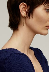 Pendientes en tono dorado que presentan un pendiente de piedra cuadrada y un largo colgante tipo cadena, colocados en un modelo que lleva un suéter azul marino.