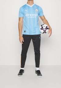 Maglia da calcio azzurro chiaro con strisce verticali, logo "ETIHAD AIRWAYS", abbinata a pantaloni sportivi neri con strisce laterali arancioni. Il giocatore tiene una palla da calcio con motivo a fantasia.