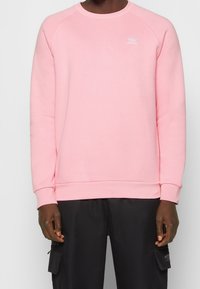 Vaaleanpunainen collegepaita, jossa on pyöreä kaula-aukko, raglan-hihat, resorit hihoissa ja brodeerattu logo rinnassa. Kangas näyttää pehmeältä ja teksturoidulta.