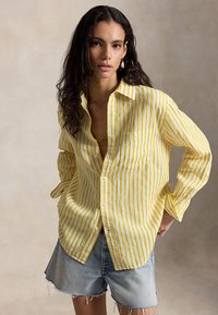 Kvinne med langt mørkt hår som har på seg en gul og hvit vertikalt stripete skjorte, oppkneppet øverst, og lyse denimshorts med frynsete kant.