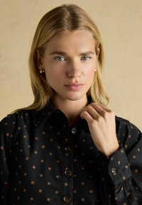 Melna krekls ar zelta zvaigžņu rakstu, ar pilnu pogu aizdari un aprocēm ar gumiju. Iezīmēts ar apkakli un teksturētu audumu.