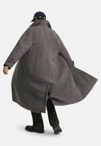 Persoon die een lange, bruine houndstooth-jas en zwarte schoenen draagt, halverwege een draai, met de kraag van de jas omhoog en een zwarte baseballcap met vlagpatch.