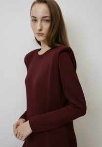 Burgundska haljina dugih rukava s podstavom na ramenima, okruglim izrezom i glatkom teksturom. Jednostavan dizajn, prilagođena silueta, minimalni šavovi.