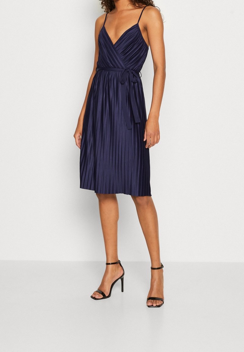 Femme portant une robe portefeuille plissée sans manches bleu marine avec fines bretelles et des sandales à talons hauts noires sur un fond clair uni.