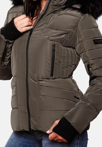 Vrouw in een bruine gewatteerde winterjas met een zwarte rits en geribbelde manchetten, haar hand die de kraag aanpast en leunend op de heup.