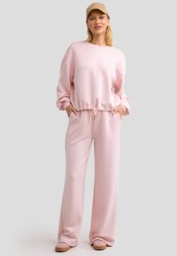 Conjunto de sudadera rosa claro con un suéter de corte holgado que presenta una cintura ajustada y pantalones relajados a juego, ambos en tela suave; incluye una gorra beige.