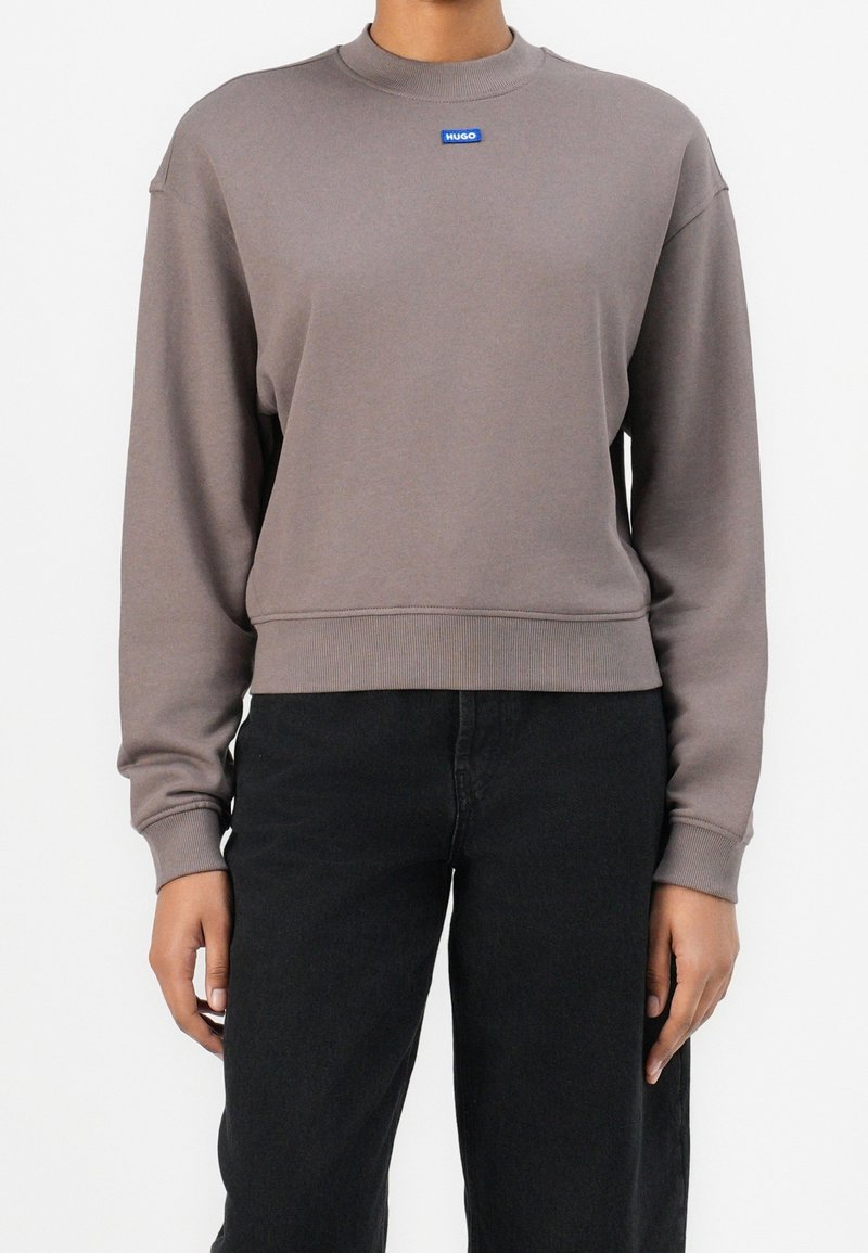 Personne portant un sweat-shirt taupe à manches longues avec un petit logo bleu « HUGO » sur la poitrine et un pantalon noir sur un fond uni.