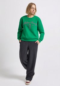 Zelená mikina s písmem a emblémem, doplněná tmavě šedými širokými kalhotami. Neformální látka s žebrovanými manžetami a pasem.