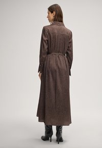 Abito lungo marrone con motivo a croco testurizzato, maniche con polsini a bottone e vita con cintura. La modella indossa stivali neri con tacco alto.