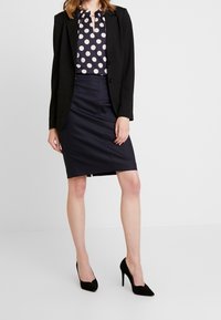 Svart kavaj över en marint blå blus med polka prickar och nyckelhålskant; matchad med en figurnära marinblå blyert kjol och svarta högklackade skor.