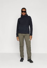 Kvinde stående iført sort bluse med lange ærmer, olivengrønne outdoor-bukser, sorte sko og store sorte solbriller mod ensfarvet baggrund.