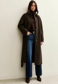 Donkerbruine lange jas met knoopsluiting, gedragen over een bruine coltrui en brede, donkere jeans. Heeft een gouden riem en puntige, bruine schoenen.