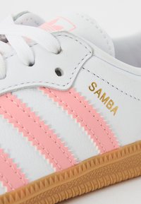 White Adidas Samba sneaker with pink stripes, gold "SAMBA" text, white laces, and a gum rubber sole.
