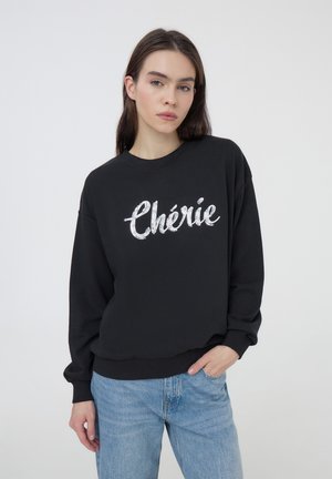 Felpa nera con la scritta "Chérie" in paillettes argentate. Presenta un col rotondo e maniche lunghe, abbinata a jeans azzurri chiari.