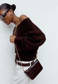 Rjav pulover iz pletenine z napihnjenimi rokavi, združen z belimi hlačami in majhno rjavo torbico z vtisnjenim krokodiljim vzorcem, verižnim pasom in okrasnimi žeblji.