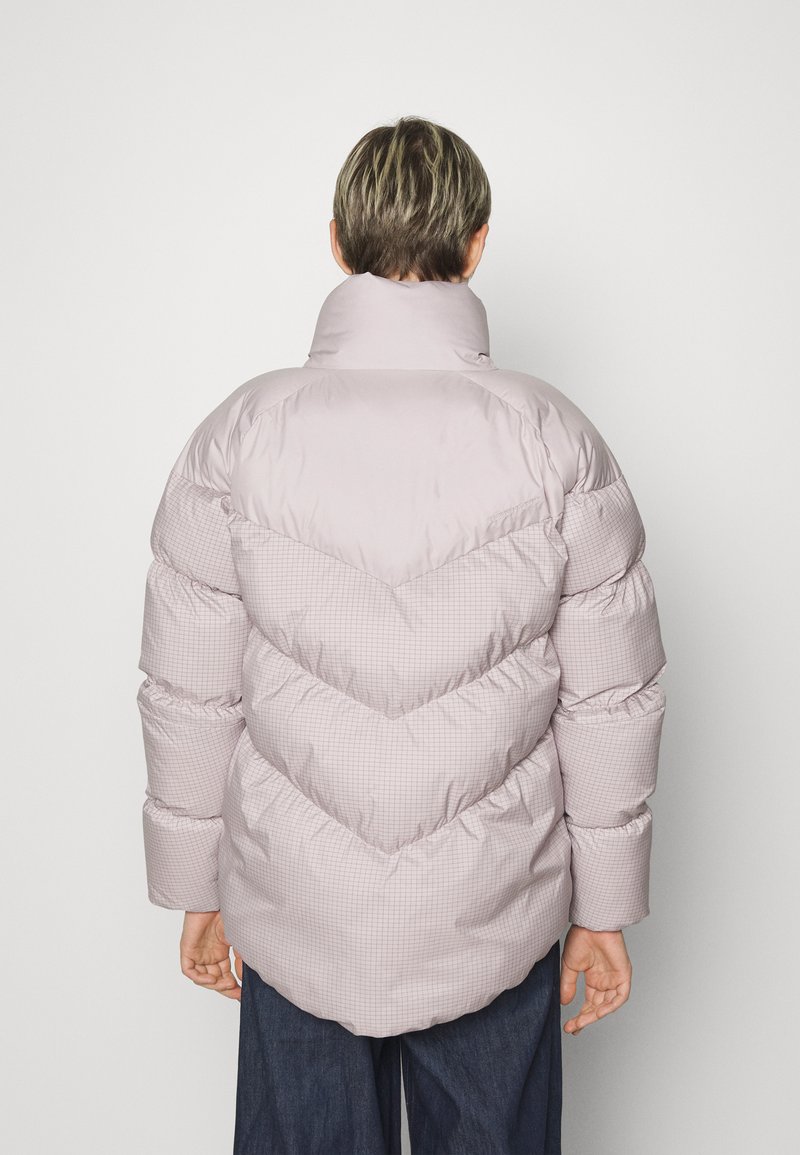 Veste en peluche rose clair avec un motif en grille texturé, un col haut et des coutures en chevron. Présente des manches larges et rembourrées ainsi qu'une coupe décontractée.
