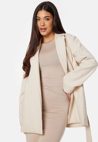 Cardigan aperto color crema realizzato in un tessuto morbido, con maniche lunghe e vestibilità rilassata, indossato sopra un vestito aderente beige.
