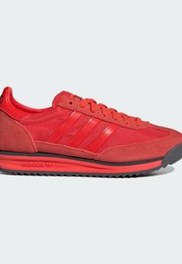 Zapatilla deportiva roja con parte superior de ante y material sintético, que presenta tres rayas blancas, una suela texturizada y un diseño de punta redondeada.