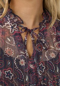 Transparante, gemuste blouse met bloemenpatronen in marineblauw, bordeaux en roomkleur. Heeft een gegolfde halslijn en een strik bij de kraag. Zwarte knoopdetails.