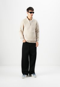 Beige fleece trøje med halv lynlås fortil, parret med løse sorte bukser og blå og hvite sneakers. Modellen bærer mørke solbriller.