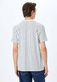 Camisa de manga corta en blanco con rayas verticales oscuras, hecha de tejido texturizado. Presenta un cuello redondo y un ajuste relajado.