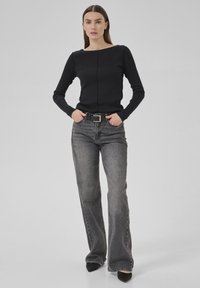 Woman wearing black long-sleeve top, faded gray straight-leg jeans with black belt, and black pointed shoes, standing with hands in pockets.