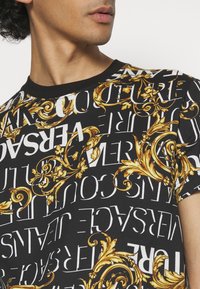 Svart kortärmad t-shirt med djärv vit text och gyllene barockinspirerade detaljer. Rund nacke med en slät yta.