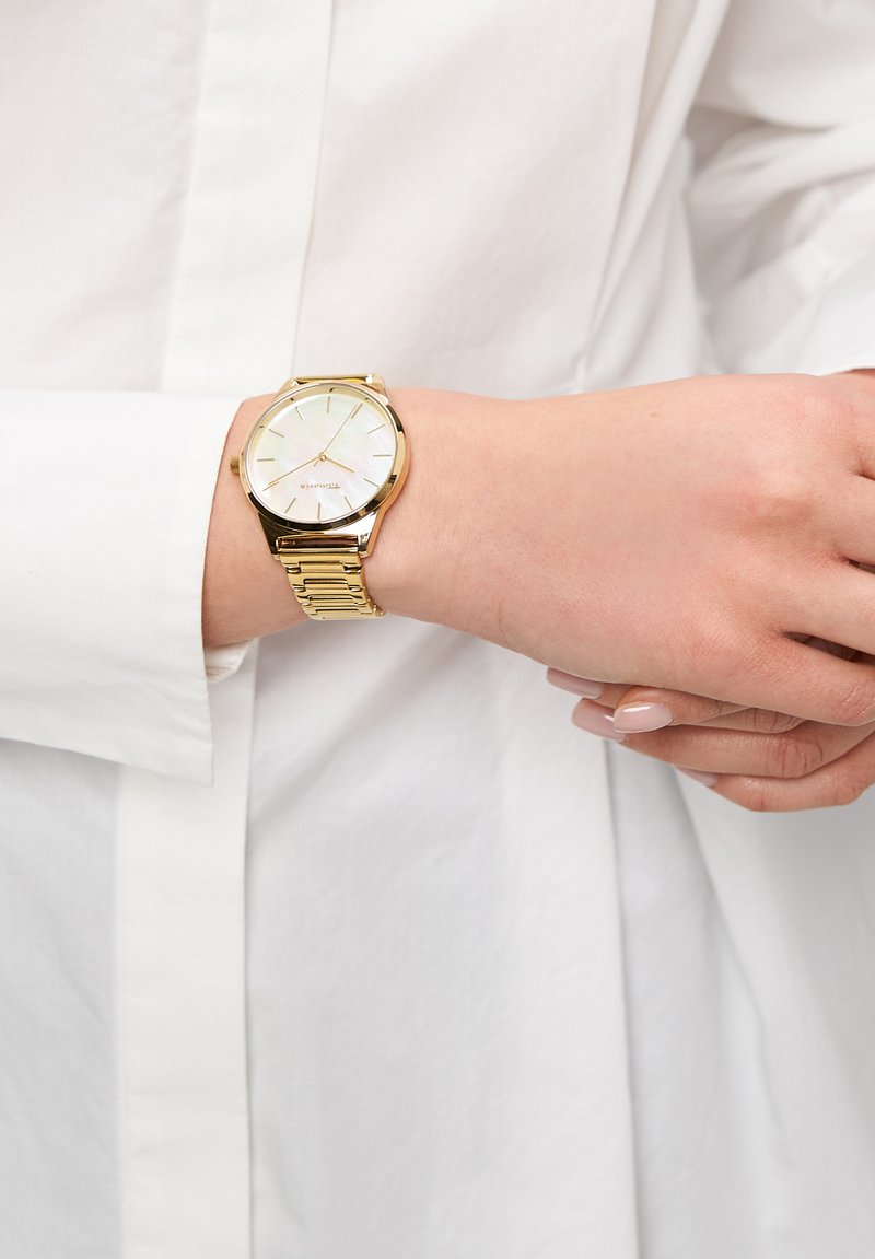 Goldfarbene Metallarmbanduhr mit runder Gehäuseform, weißem Zifferblatt und schlanken Zeigern. Sie verfügt über ein strukturiertes Armband und dezente Logodetails.