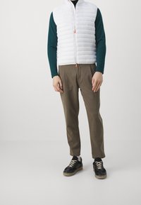 Hvid puffer vest med horisontale syninger, orange lynlås træk, grøn langærmet skjorte, brune bukser, og sorte sneakers med gummisål.