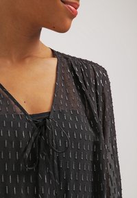 Blusa negra transparente con acentos metálicos verticales, escote en forma de v, detalle de lazo en el frente y tejido texturizado. Ideal para superponer.
