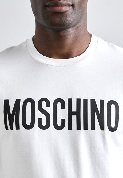 Tricou din bumbac alb, cu un imprimeu îndrăzneț „MOSCHINO” de culoare neagră. Mâneci scurte și un design cu guler rotund. Textură netedă.