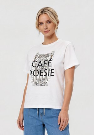 Lyshåret kvinde iført en hvid T-shirt med sort arkitektonisk tryk og teksten "CAFÉ POÉSIE", kombineret med blå jeans på en ensfarvet baggrund.
