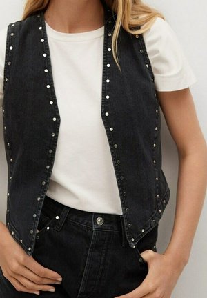 Frau trägt ein weißes Kurzarmhemd unter einer schwarzen ärmellosen Jeansweste mit silbernen Nieten und passenden schwarzen Jeans mit hoher Taille.