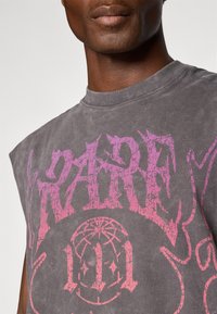 Mand iført en falmet grå ærmeløs skjorte med slidt pink grafik tekst og et cirkulært emblem på brystet.