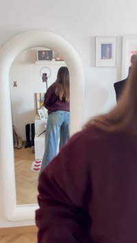 Vrouw met lang haar, gekleed in een wijnrode trui en blauwe jeans, staat voor een rondboogspiegel in een kamer met kunstwerken, laarzen en een ringlamp.