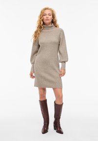 Smėlio spalvos megztinė su aukštu apykakliu, ilgos pūstos rankovės ir tiesi apačia. Dėvima kartu su rudos odos auliniais iki kelių.