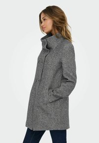 Manteau en laine gris avec un col haut, des boutons sur le devant et des poches latérales. Il possède une finition texturée et un design mi-long.