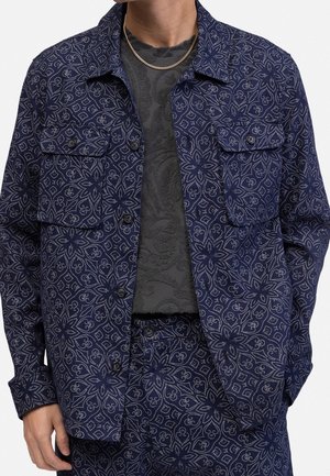 Hombre con chaqueta azul oscuro de estampado y pantalones a juego sobre una camisa negra texturizada con un collar de cadena dorada.