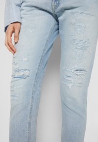 Jeans de mezclilla azul claro con detalles rasgados, bordes deshilachados y una textura descolorida. El corte es recto, con costuras visibles.