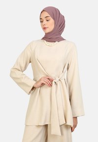 Beige long-sleeve tunic with a tied waist, made of lightweight fabric. Features a round neckline and a smooth texture. Accented with a pearl necklace.