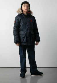 Granatowa kurtka puchowa z kapturem wykończonym futrem, pomarańczowymi zamkami błyskawicznymi i przednimi kieszeniami. W komplecie z ciemnymi spodniami cargo i czarnymi sneakersami.