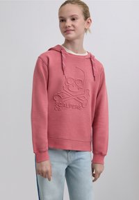 Pink hoodie made of soft fabric, featuring a raised skull and crossbones design. It has a drawstring hood and ribbed cuffs. Worn with light blue jeans.