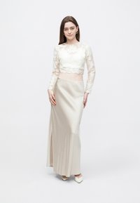 Pitsinen lyhyt yläosa, jossa pitkät hihat ja aaltoileva helma, yhdistetty beigeen satiinihameeseen, jossa on sivuhalkaisija. Tyylikkäät kengät viimeistelevät asun.