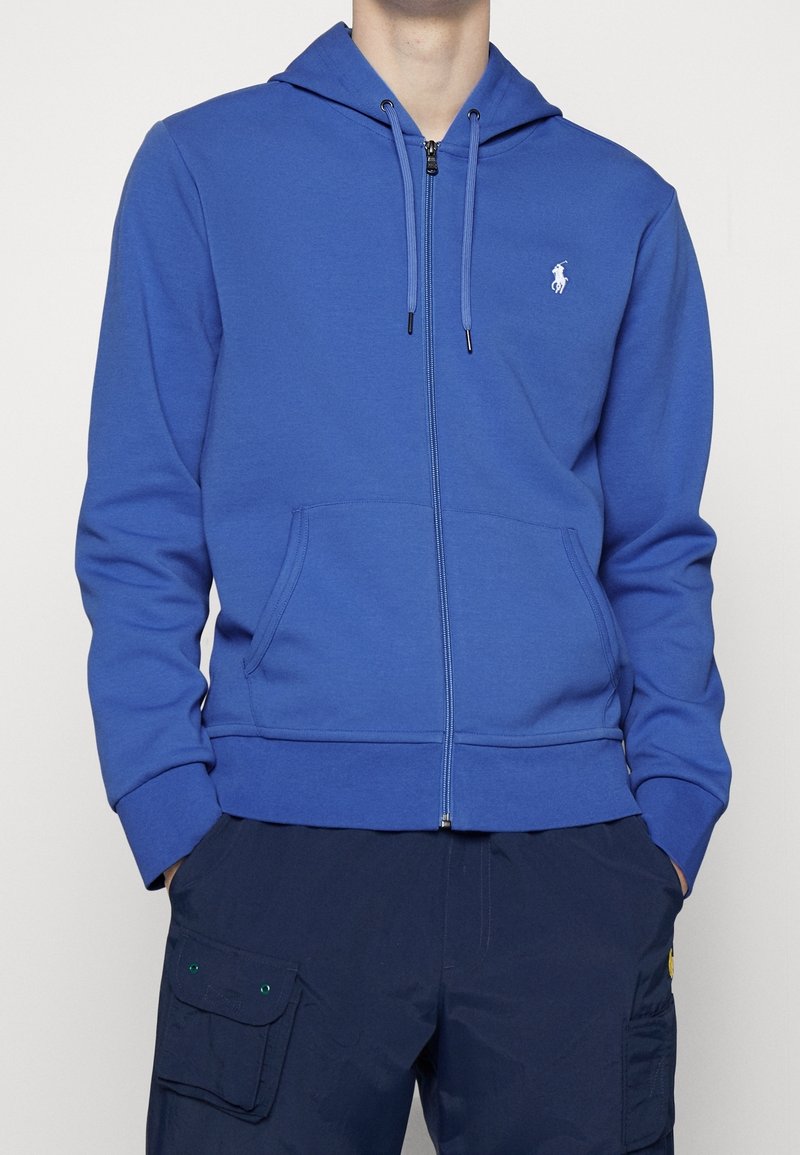 Homme portant un sweat à capuche bleu avec fermeture éclair et poches avant, ainsi qu'un pantalon cargo bleu marine avec poches à rabat, les mains dans les poches, debout devant un fond uni.